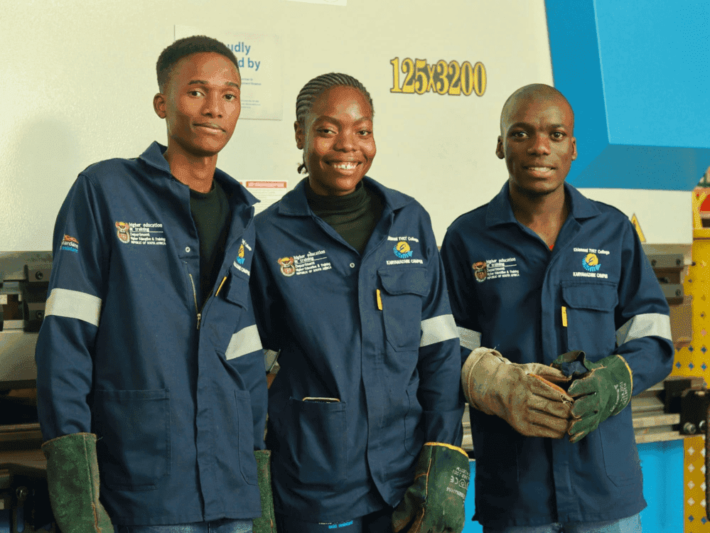 Three Ehlanzeni TVET College students in Kanyamazane Campus overalls and welding gloves, standing in a college workshop.